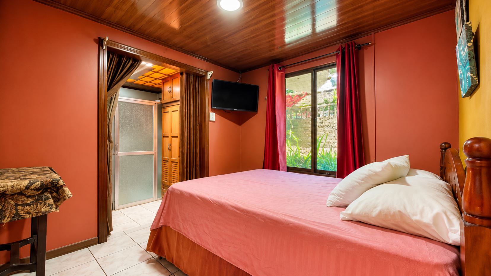 Bedroom with coral walls, a pink bedspread, white pillows, a wall-mounted TV, and a window with red curtains.