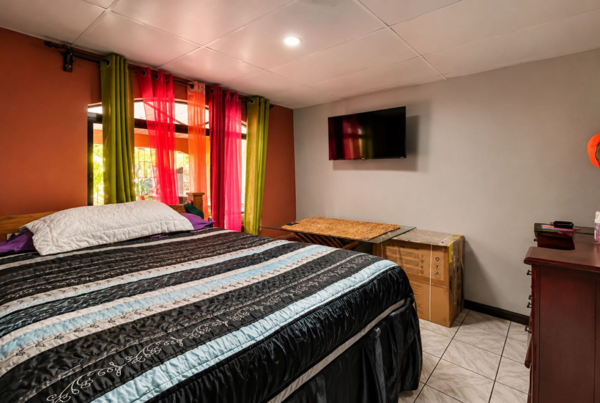 Bedroom with a large bed covered by a striped quilt, colorful sheer curtains, and a wall-mounted TV on a gray wall behind the bed's footboard