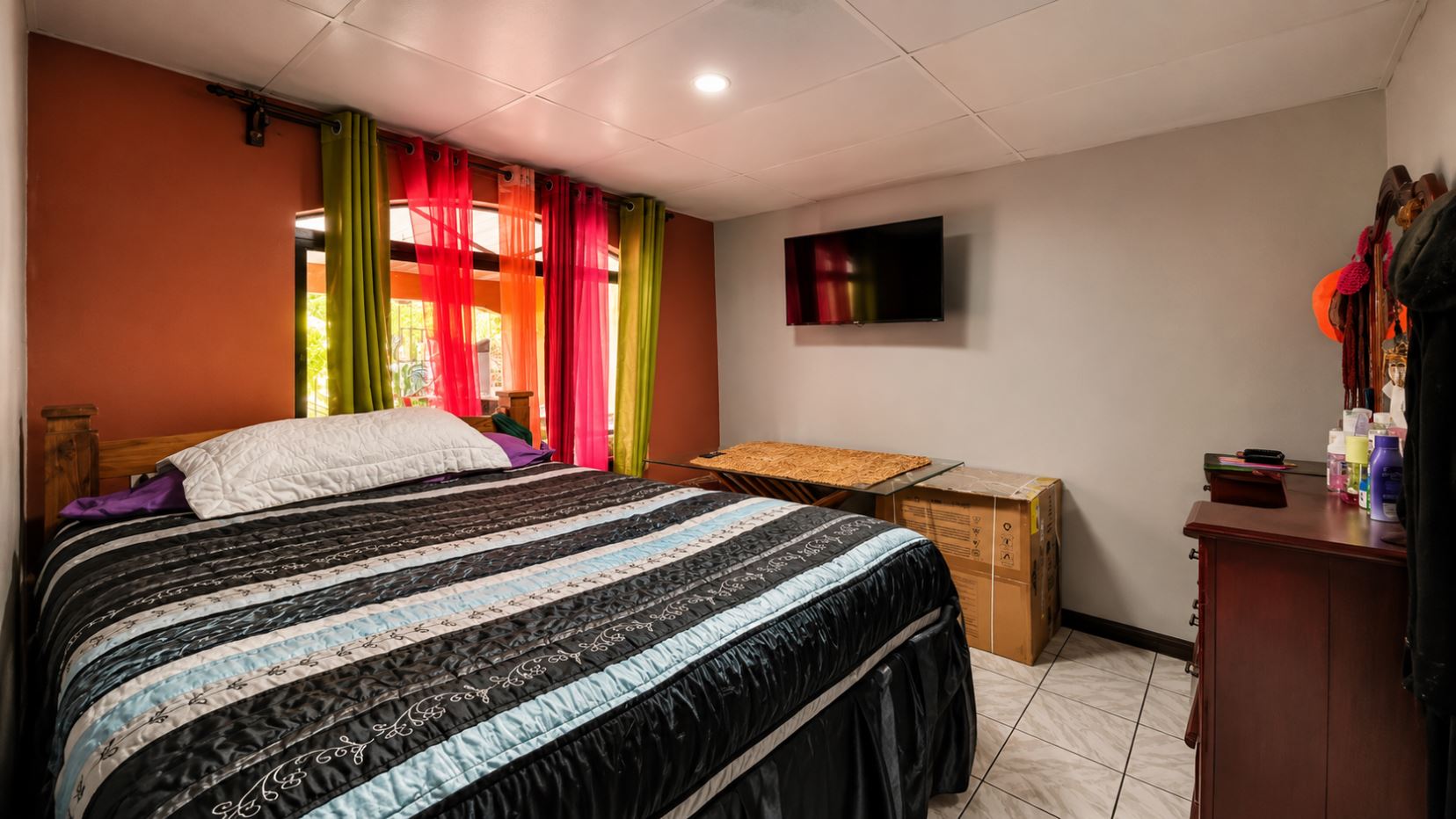 Bedroom with a large bed covered by a striped quilt, colorful sheer curtains, and a wall-mounted TV on a gray wall behind the bed's footboard