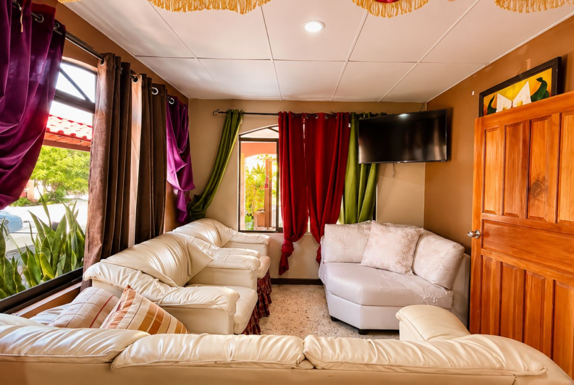 Cozy living room with white leather sectional sofa arrangement, multicolored curtains (purple, red, green, brown), and a wall-mounted flat-screen TV. A wooden door is on the right and a speckled tile floor completes the scene.