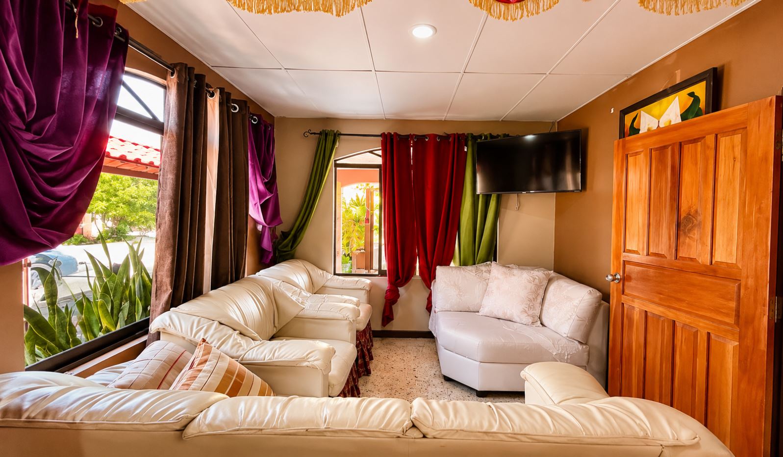 Cozy living room with white leather sectional sofa arrangement, multicolored curtains (purple, red, green, brown), and a wall-mounted flat-screen TV. A wooden door is on the right and a speckled tile floor completes the scene.