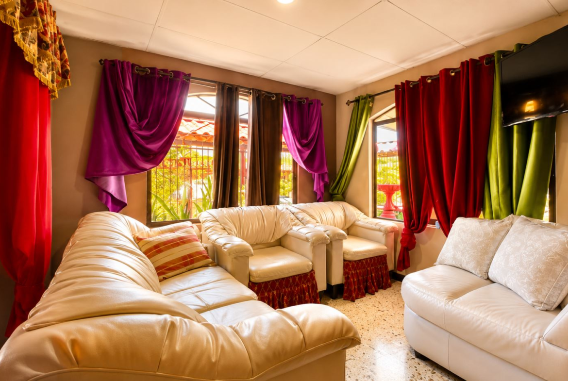 Cozy living room with white leather sectional and armchairs, colorful drapes by sunny windows, and a wall TV.
