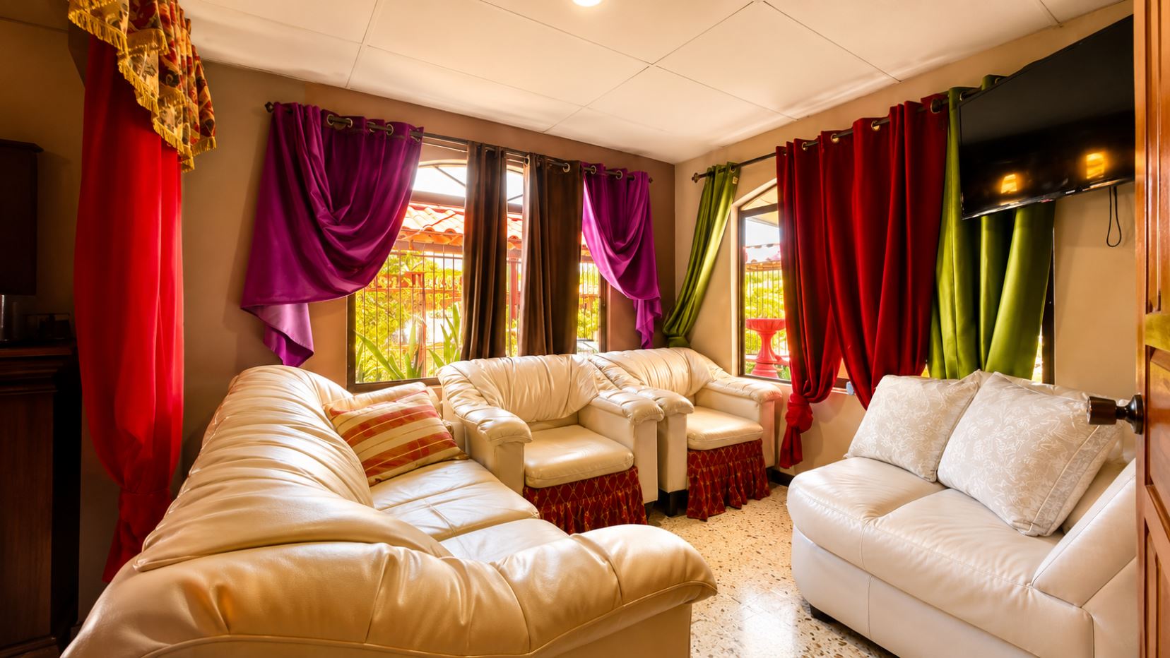 Cozy living room with white leather sectional and armchairs, colorful drapes by sunny windows, and a wall TV.