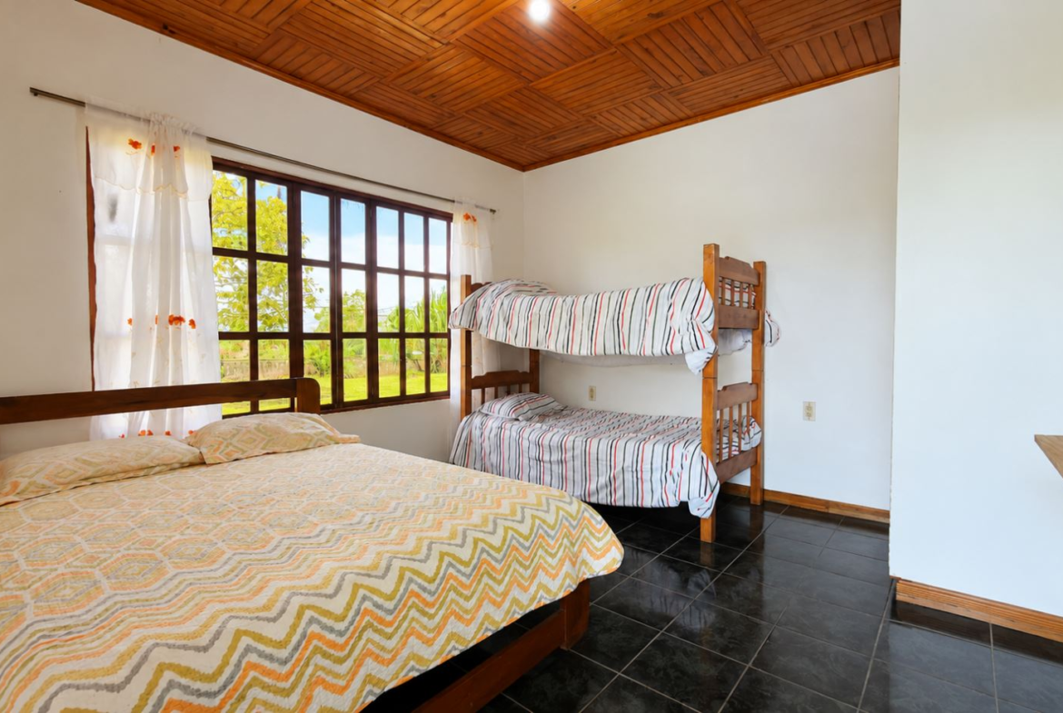 Cozy bedroom with a wooden double bed and a wooden bunk bed by a large window, white walls, and a dark tiled floor.