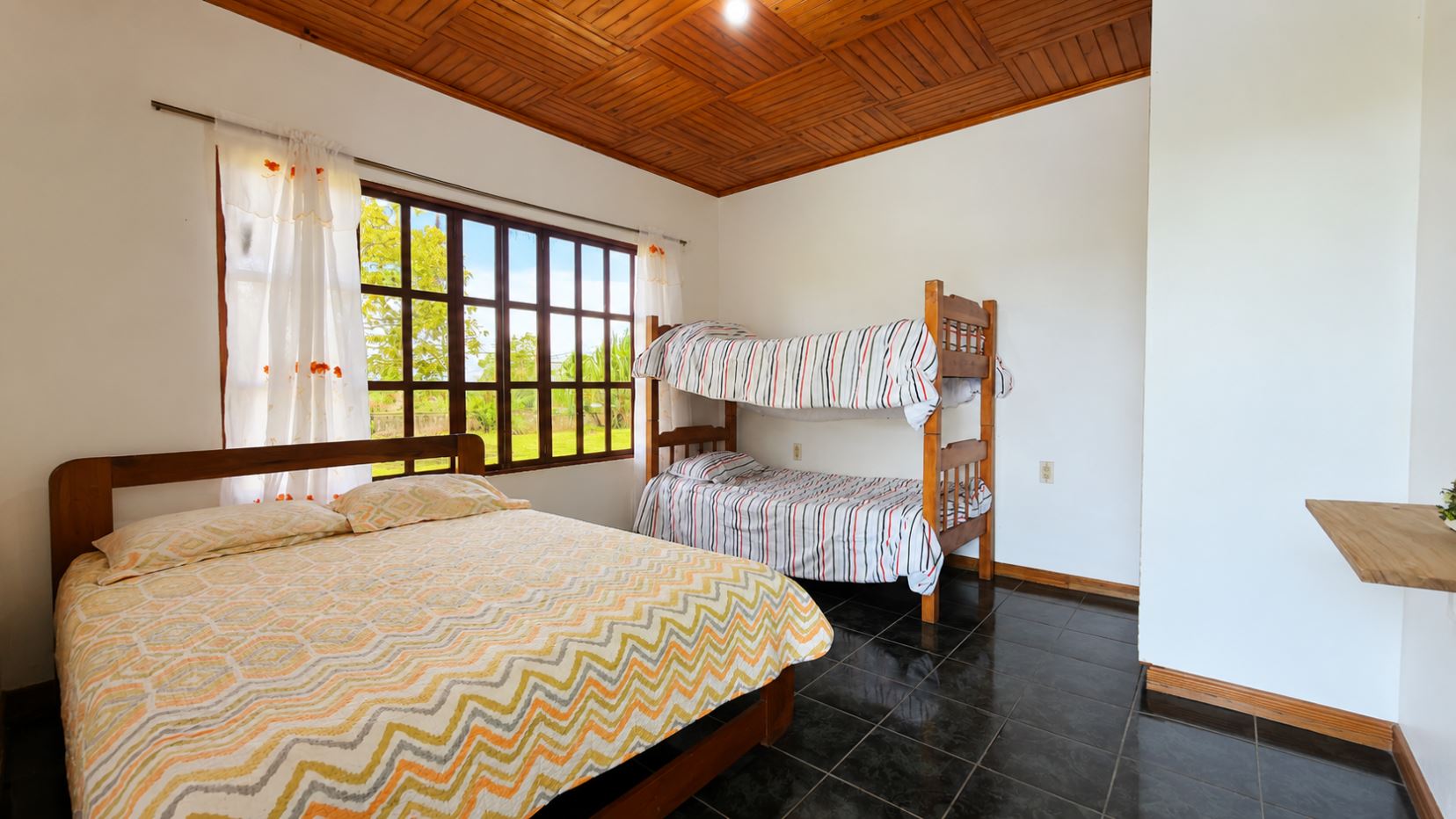 Cozy bedroom with a wooden double bed and a wooden bunk bed by a large window, white walls, and a dark tiled floor.