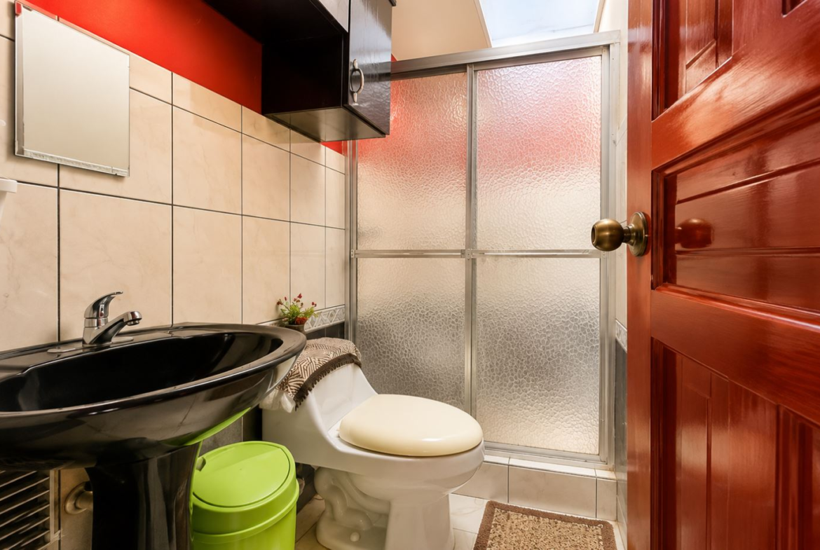 Compact bathroom: black sink, white toilet, beige tiled walls, frosted glass shower, and a wooden door.