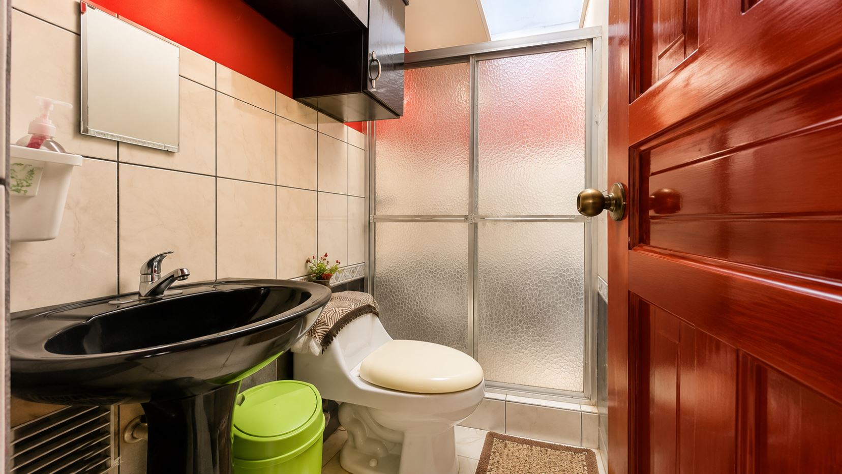 Compact bathroom: black sink, white toilet, beige tiled walls, frosted glass shower, and a wooden door.