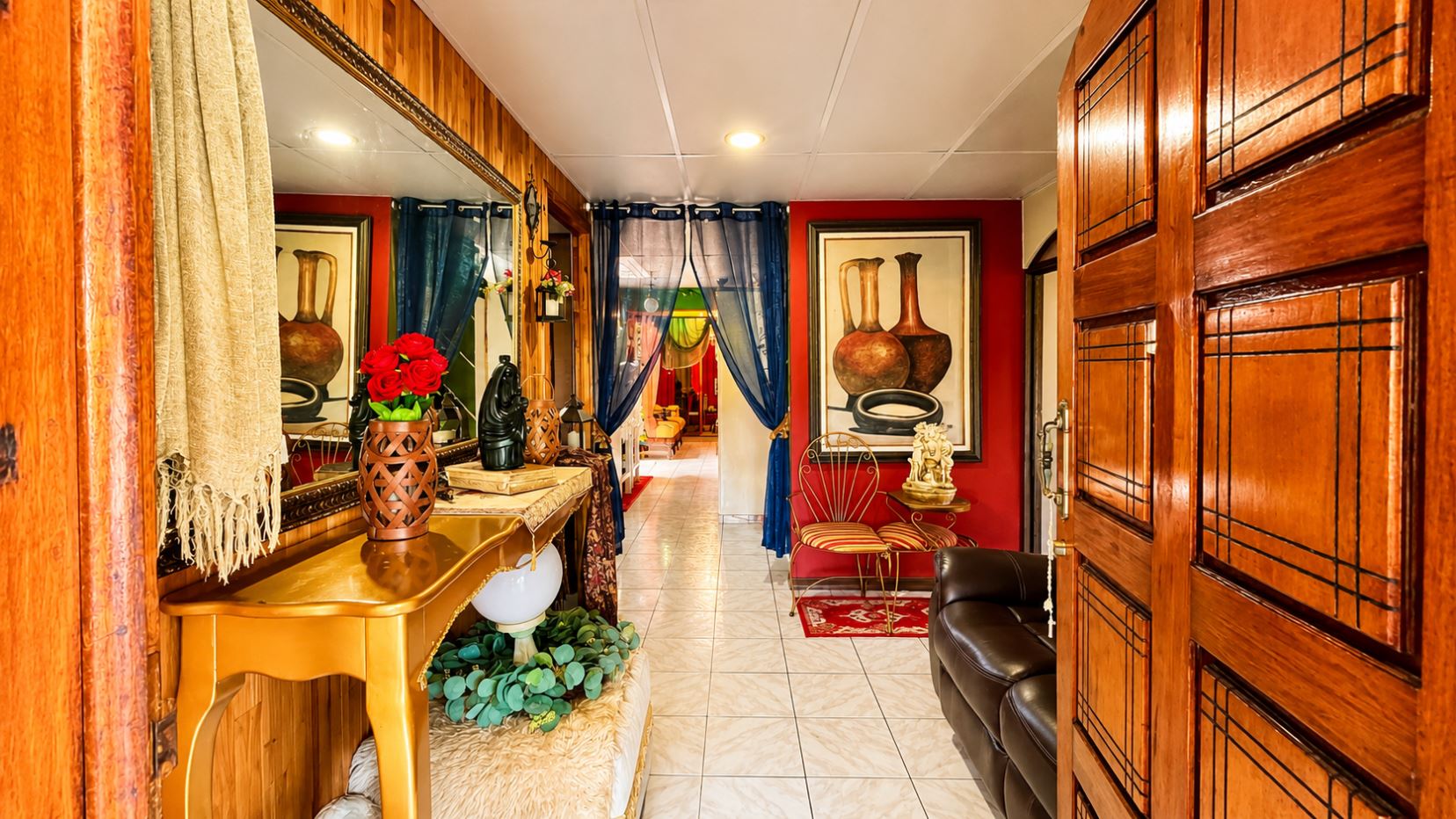 Luxurious hallway leading to a living room, with red walls, blue sheer curtains, a gold console table, and a vase of red roses.