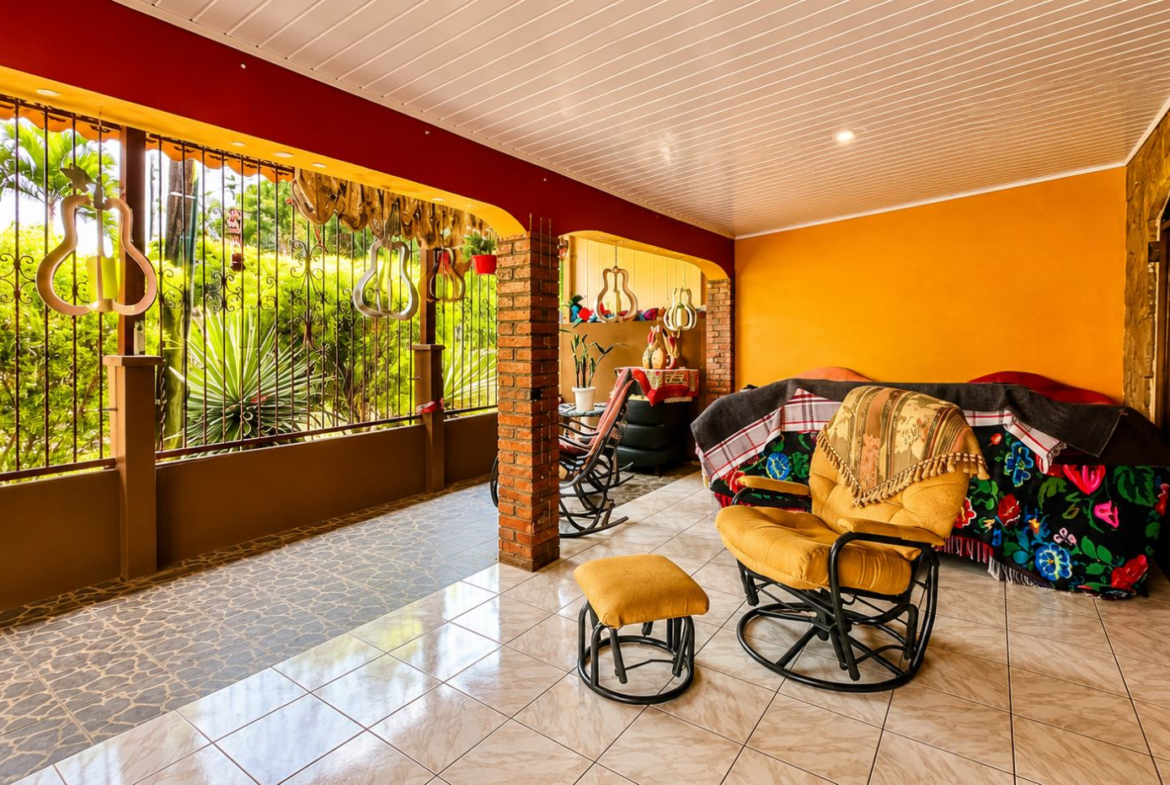 Covered patio with brick posts, orange wall, and garden view; cushioned rocking chairs and an ottoman on tiled floor, with colorful blankets.