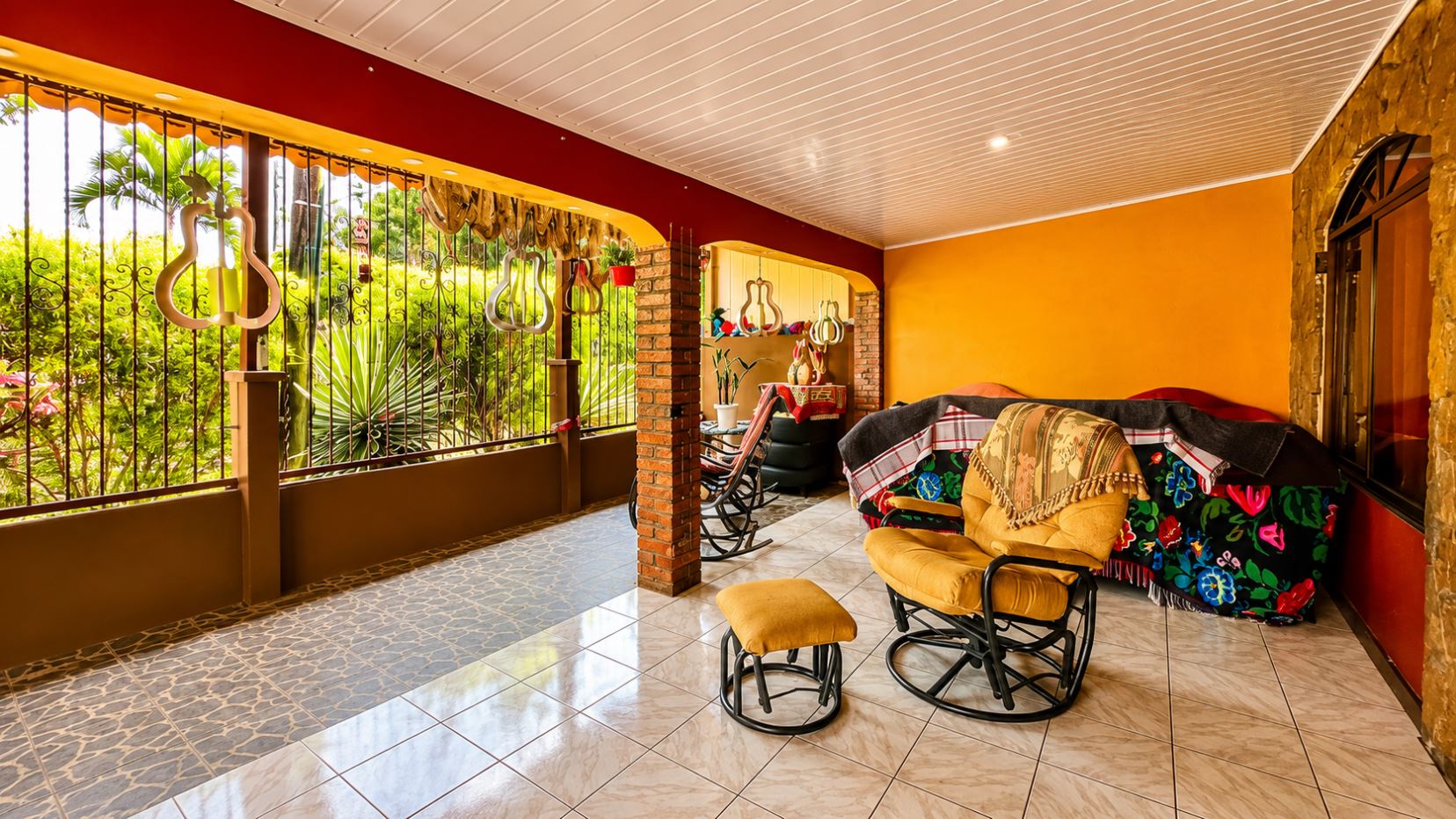Covered patio with brick posts, orange wall, and garden view; cushioned rocking chairs and an ottoman on tiled floor, with colorful blankets.