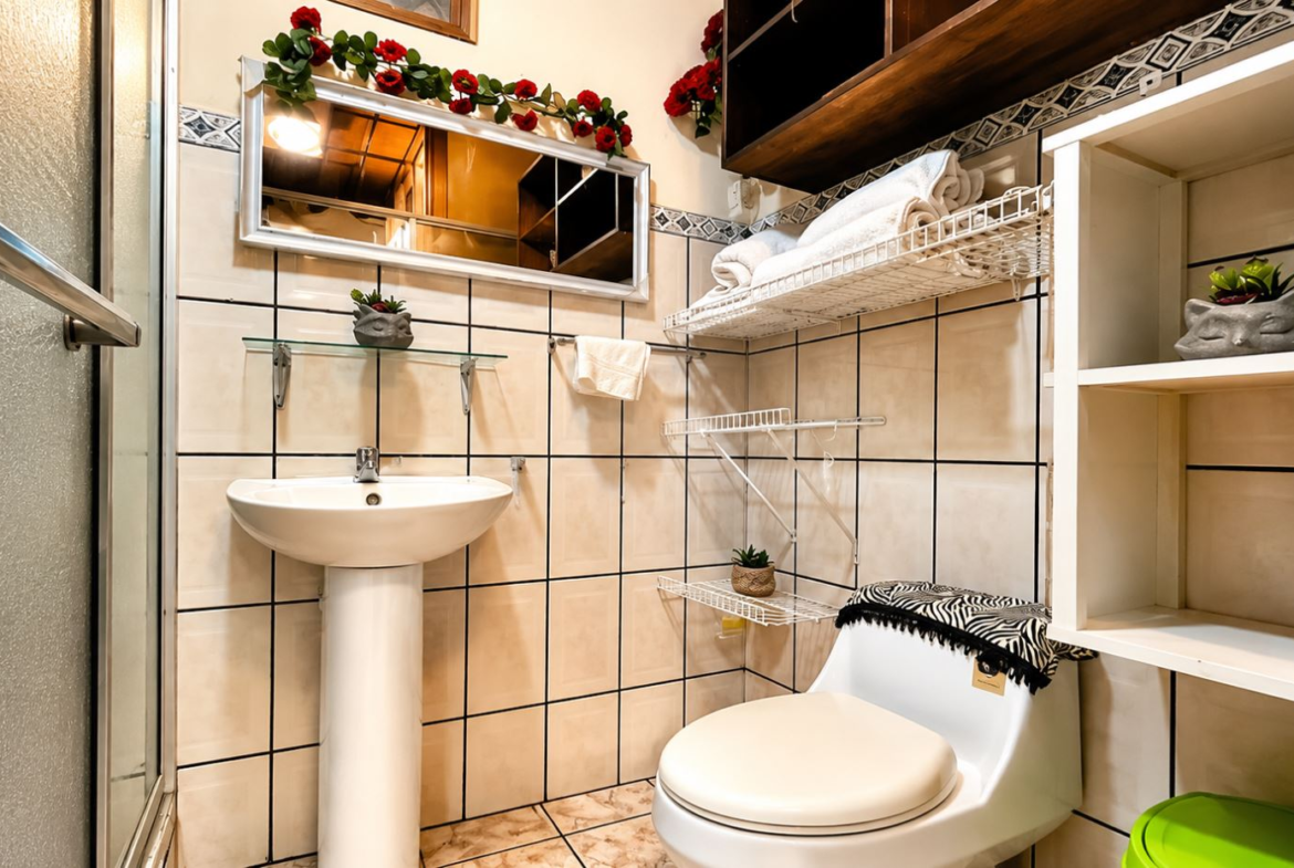 Small bathroom with pedestal sink, toilet, white shelves, and decorative red flowers above a framed mirror.