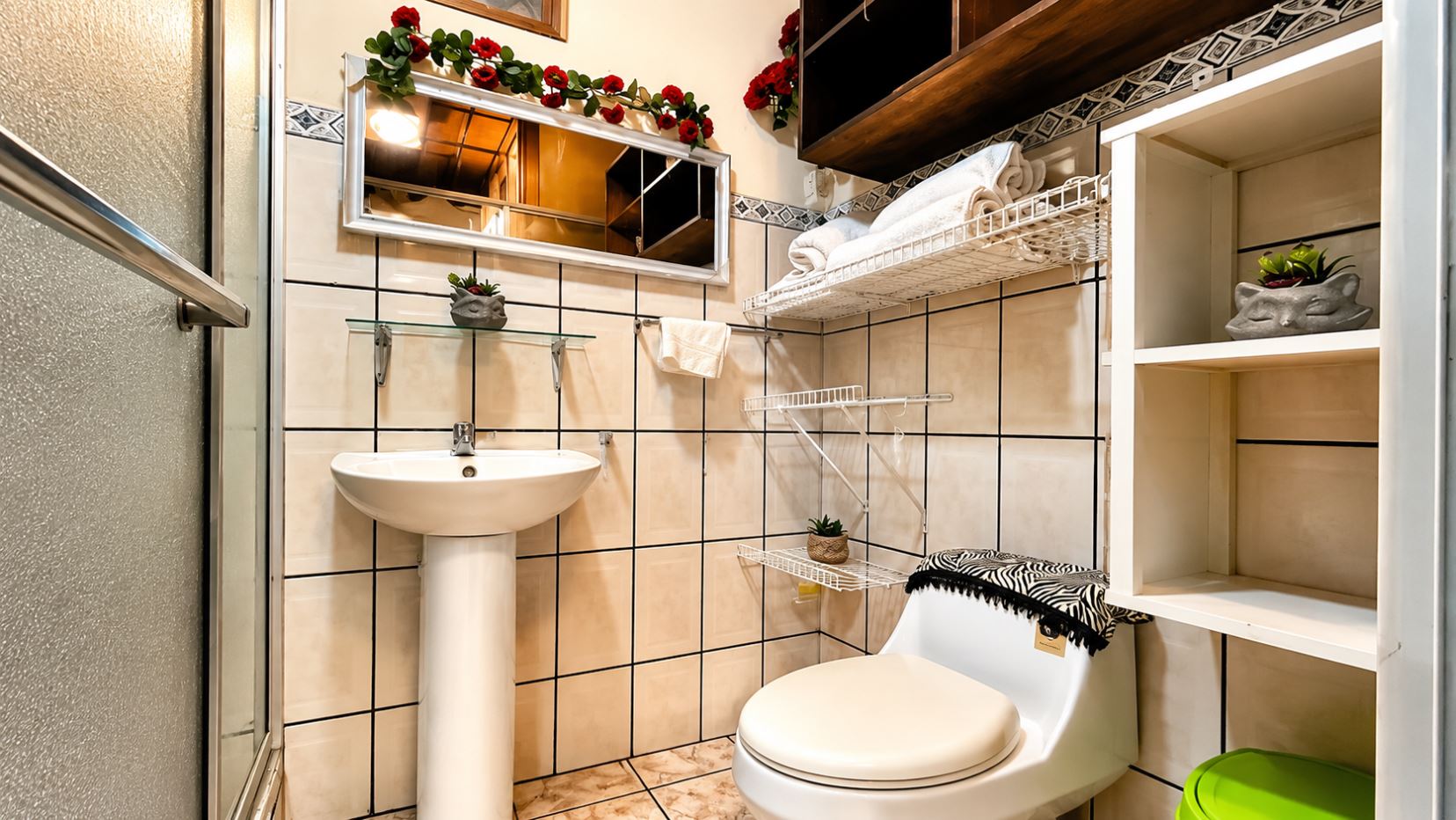 Small bathroom with pedestal sink, toilet, white shelves, and decorative red flowers above a framed mirror.