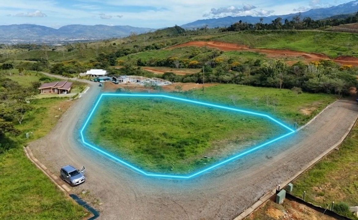 A vacant grassy plot outlined in bright blue on a curved gravel road, with green hills and a few buildings in the distance.
