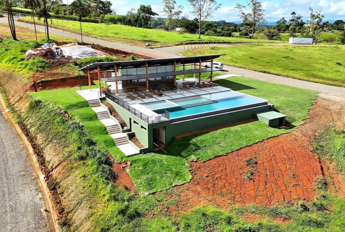 A modern hillside home with a long rectangular pool, shaded deck, and surrounding green lawns.