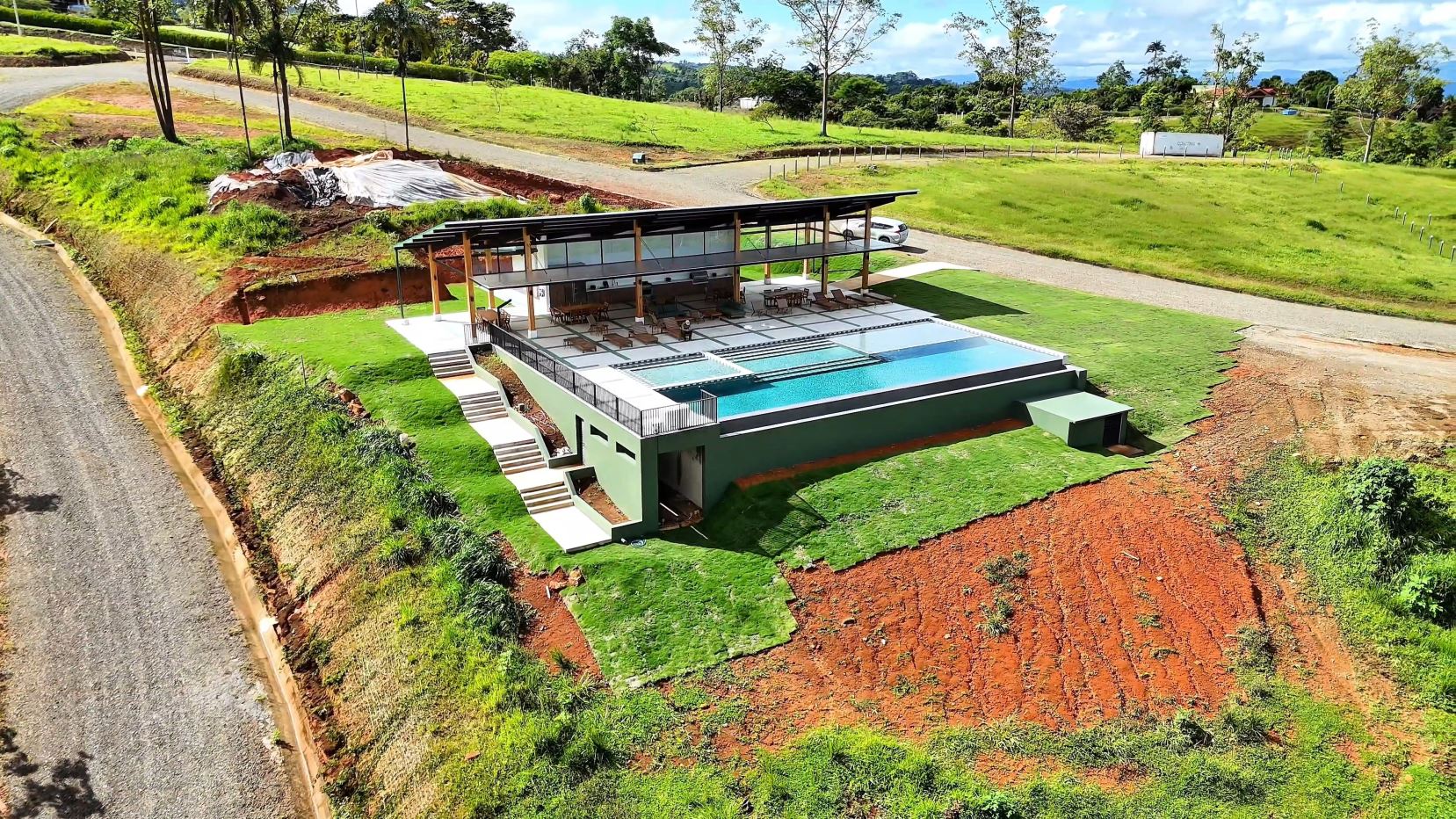 A modern hillside home with a long rectangular pool, shaded deck, and surrounding green lawns.